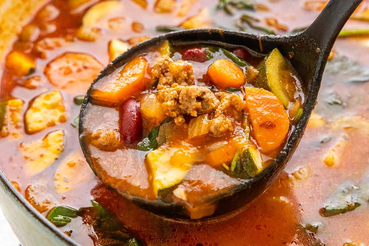 Ladle scooping Italian sausage soup with carrots, beans, zucchini, and spinach from a pot