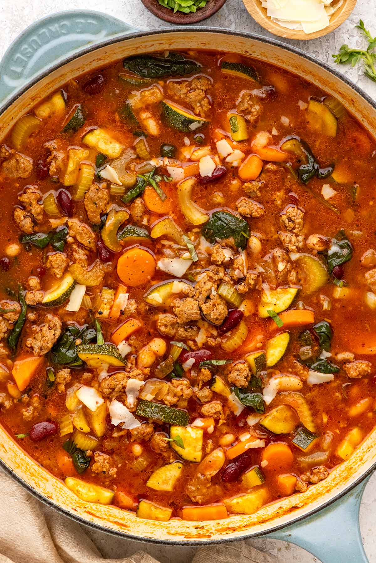 Italian sausage soup with zucchini, carrots, beans, and spinach in a large pot, topped with parmesan shavings