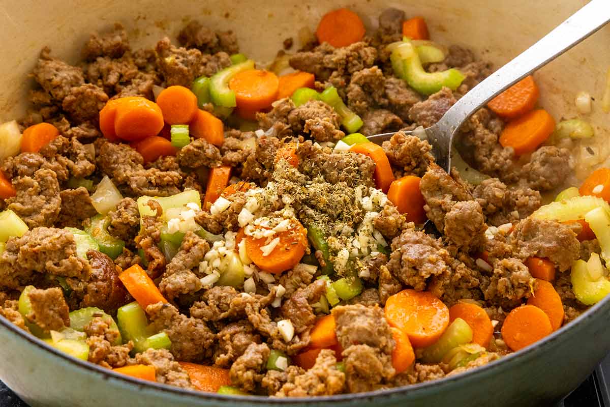 Browned Italian sausage in a pot with sliced carrots, celery, minced garlic, and dried herbs
