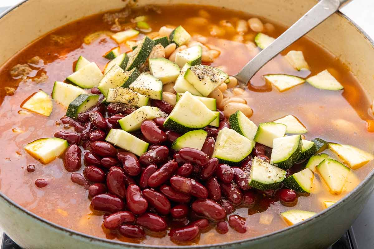 Adding red kidney beans, cannellini beans, and chopped zucchini to the soup pot with broth and sausage