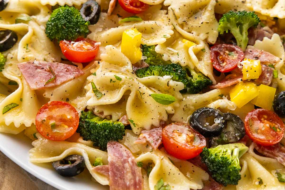 Close up of a bow tie pasta salad with pieces of salami, broccoli, tomatoes, and olives.