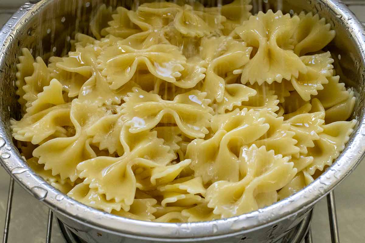 Cooked bow tie pasta in a metal colander being rinsed with water.