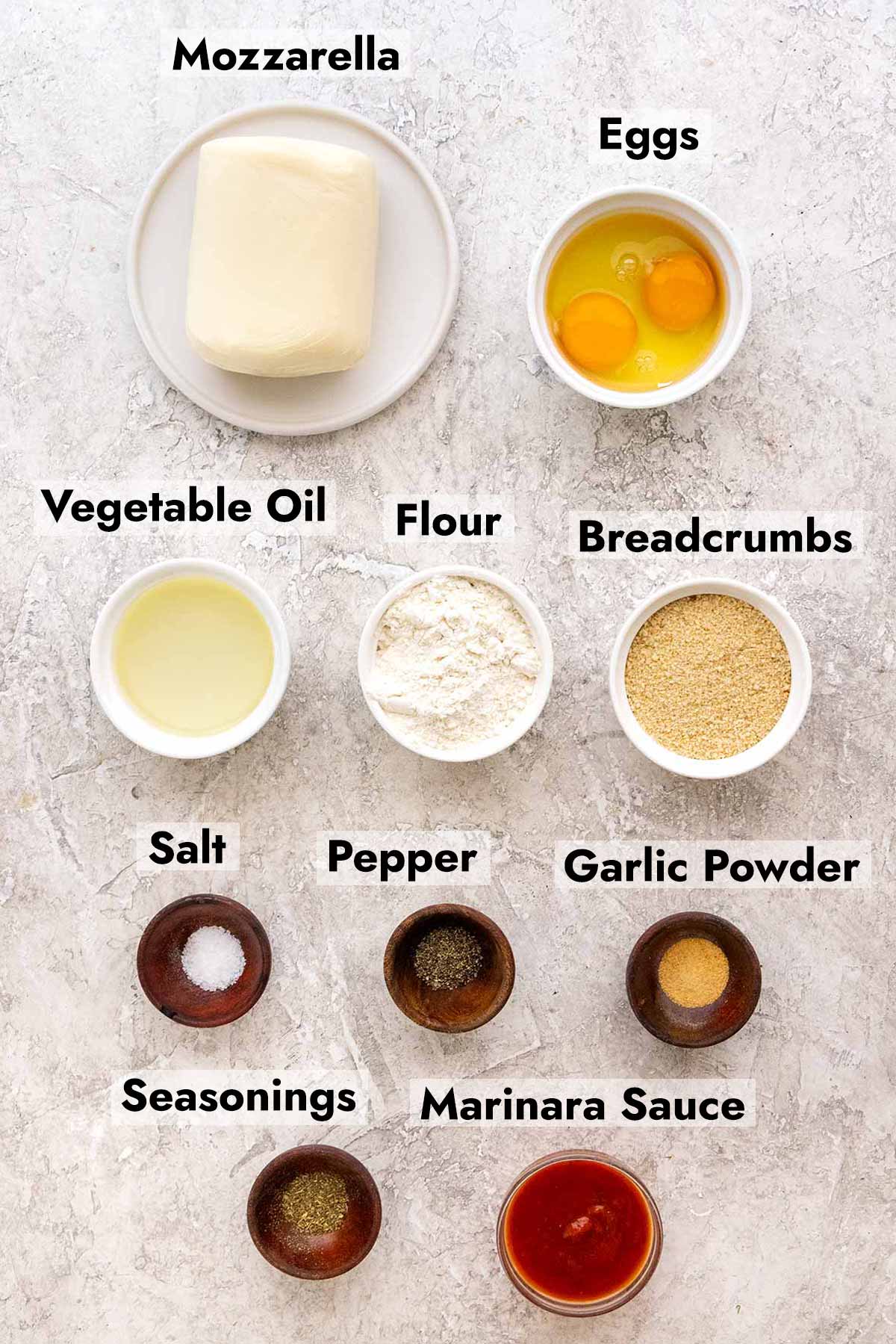 Mozzarella, eggs, oil, flour, breadcrumbs, garlic powder and other ingredients in small white bowls.