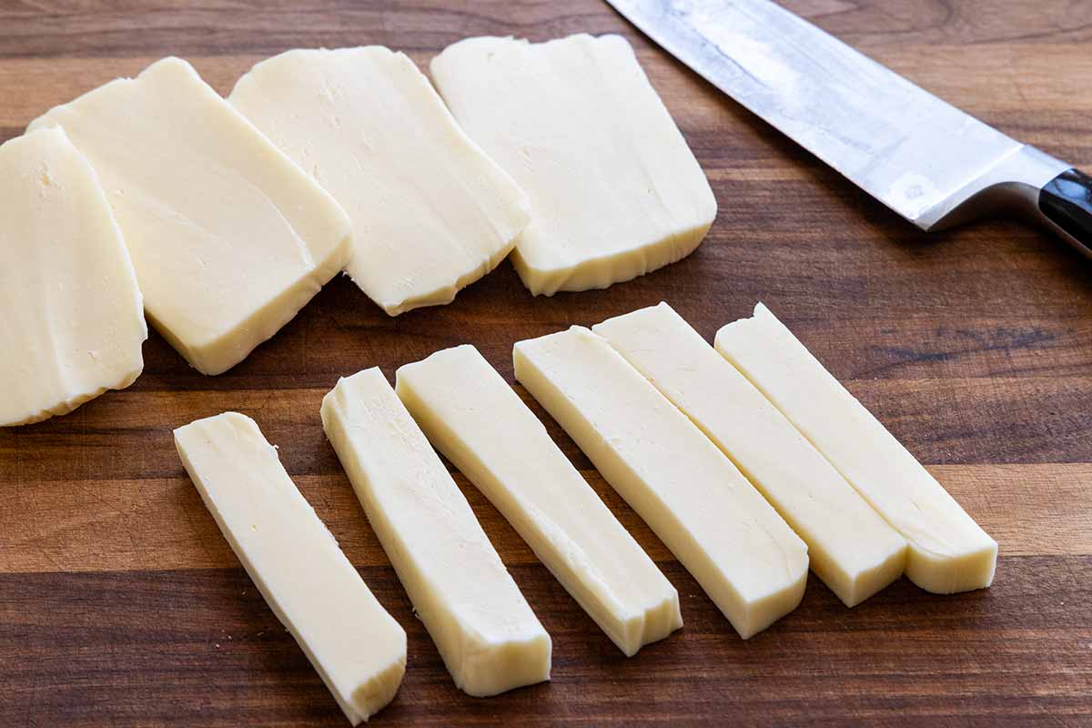 Block of mozzarella sliced into sticks on a wooden cutting board.