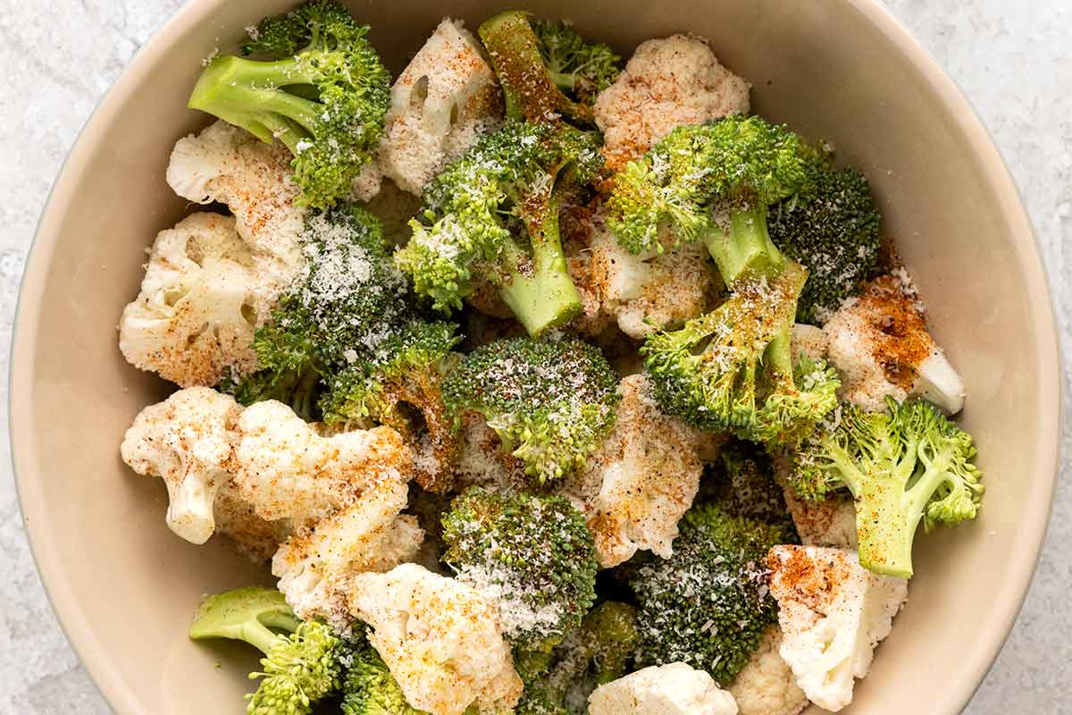 Raw broccoli and cauliflower florets in a bowl, seasoned with spices and grated Parmesan cheese