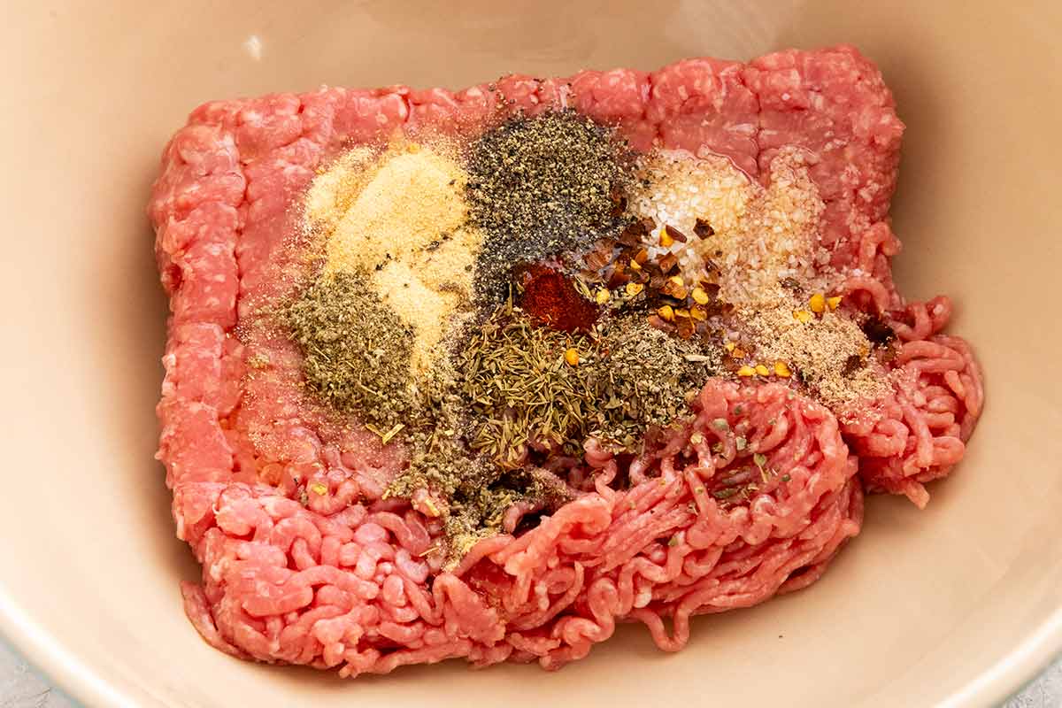 Raw ground pork in a mixing bowl topped with various dried herbs and seasonings