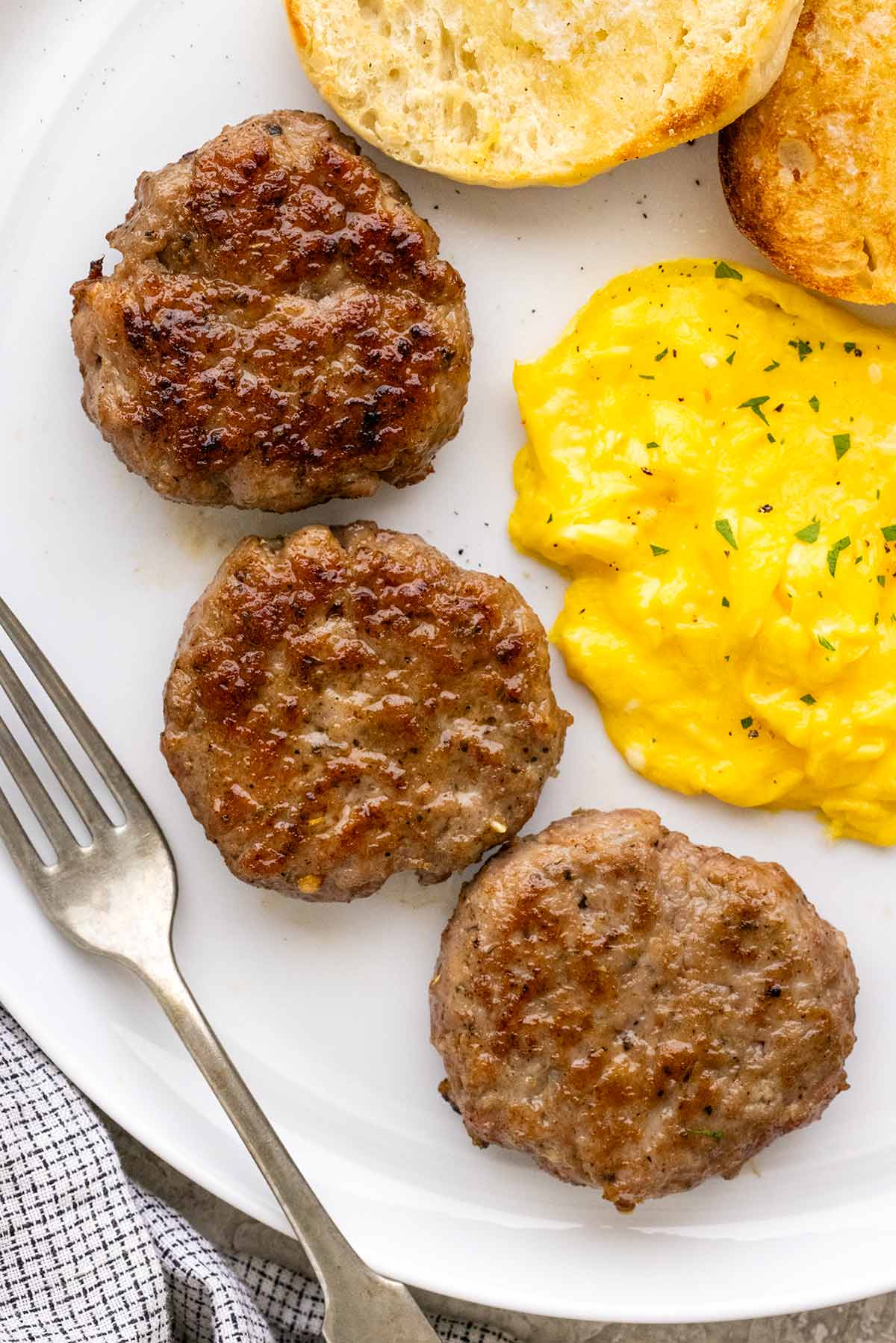 Three browned breakfast sausage patties served on a plate with scrambled eggs and toasted English muffins