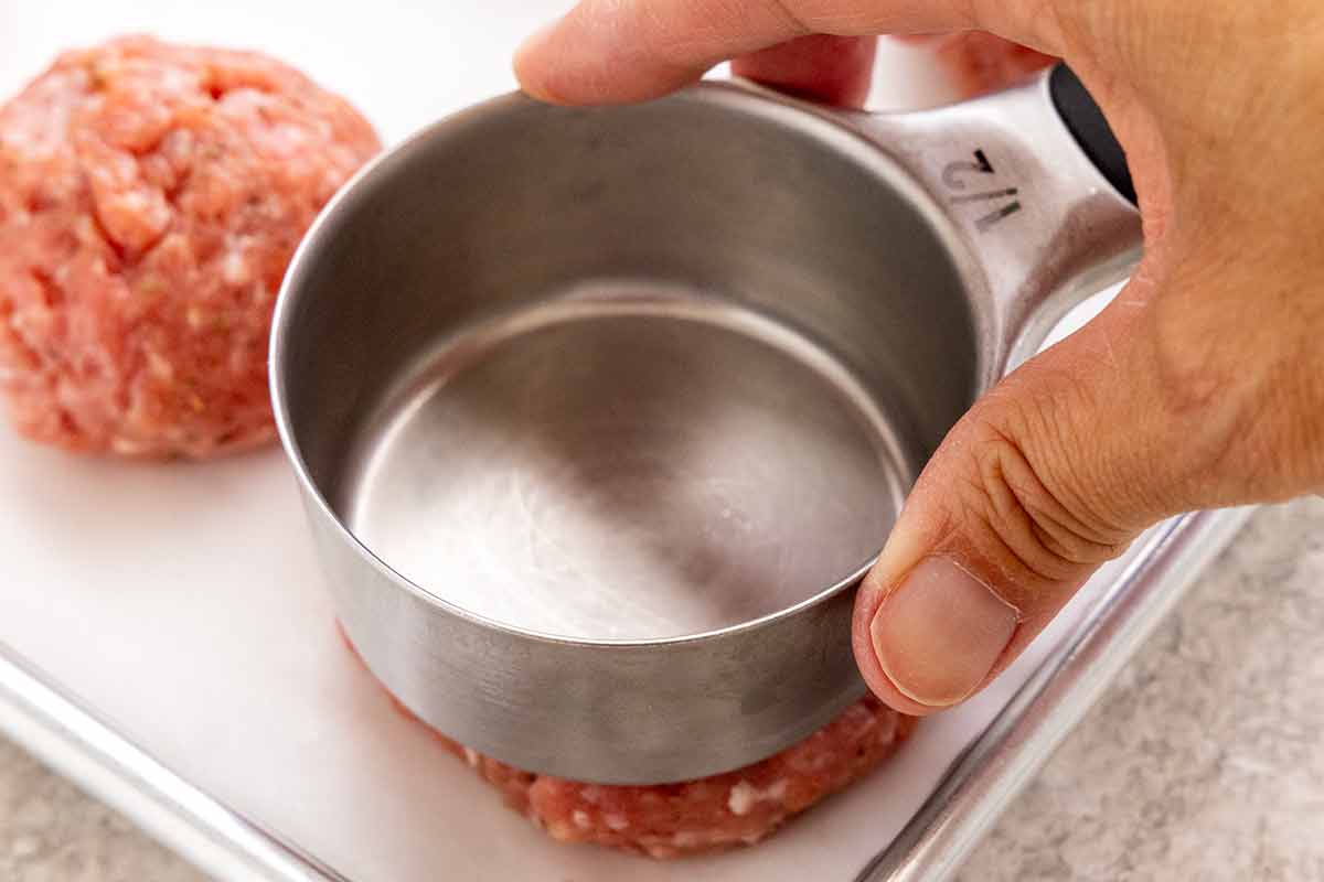 Hand using the bottom of a metal measuring cup to press a raw sausage ball into a flat patty