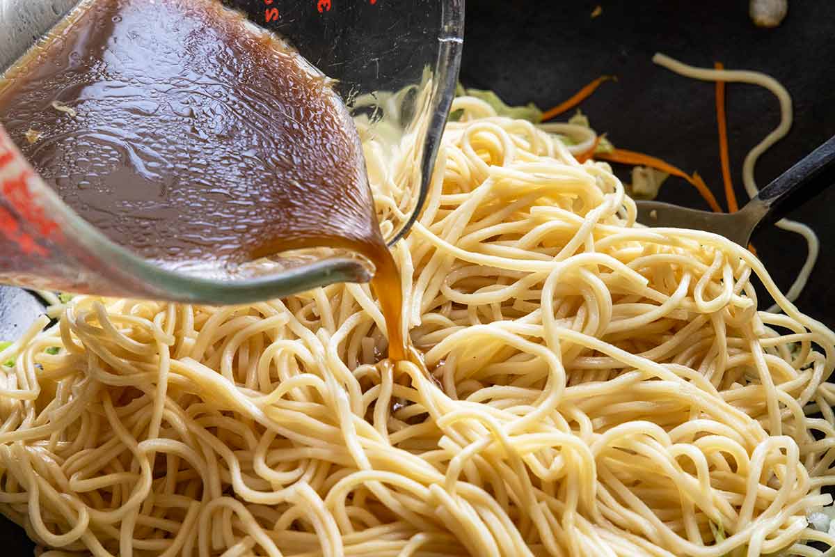Stir fry sauce being poured over lo mein noodles inside a wok.