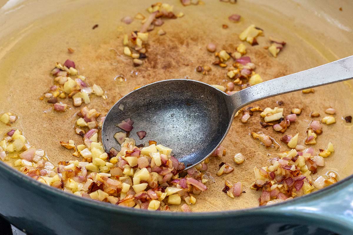 Diced red onions, garlic, and chili flakes sautéing in oil in a pan with a metal spoon.