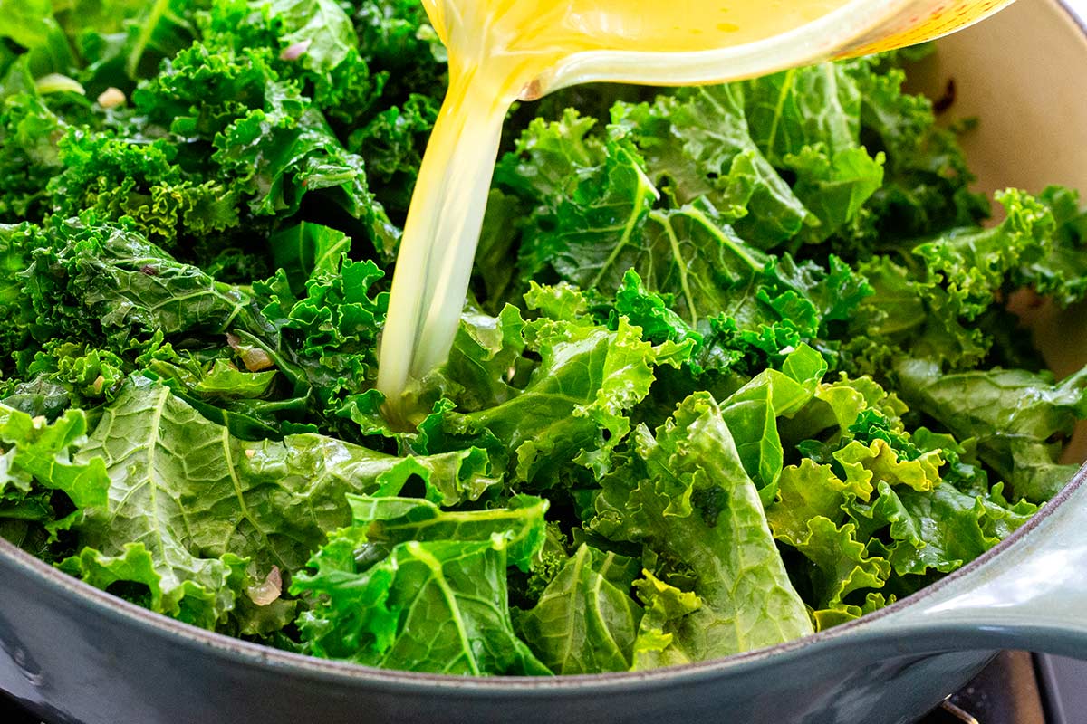 Pouring vegetable stock over chopped green kale in a pot.