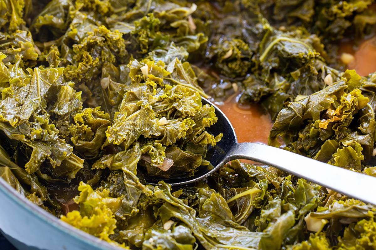 Chopped kale simmering in braising liquid inside a pot, scooped with a spoon.