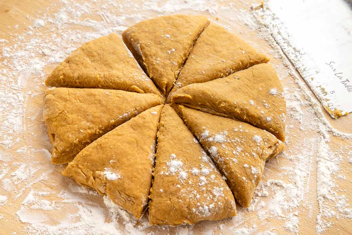 A round disk of pumpkin scone dough cut into eight triangular wedges on a floured surface.