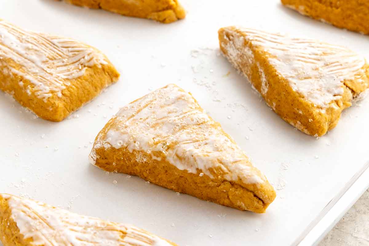 Unbaked pumpkin scone dough wedges brushed with cream and sprinkled with coarse sugar on a baking sheet.