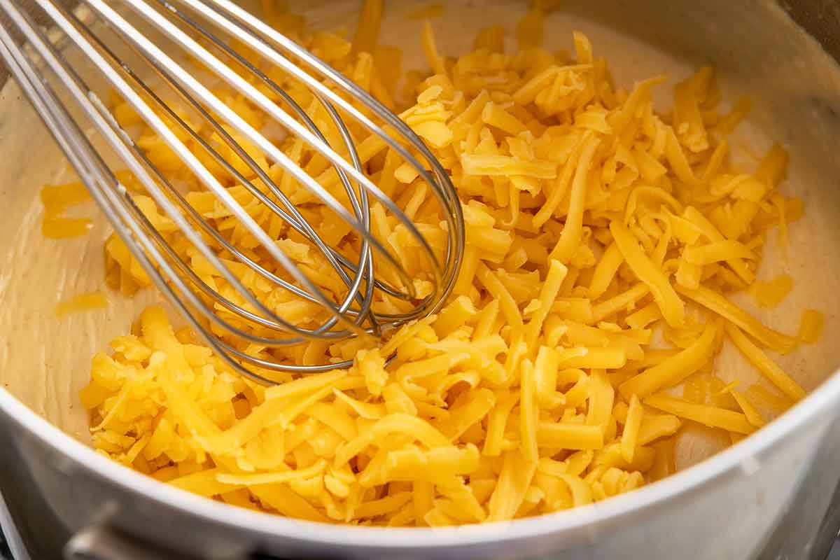 Shredded cheddar being whisked into the thickened beer sauce in a saucepan.
