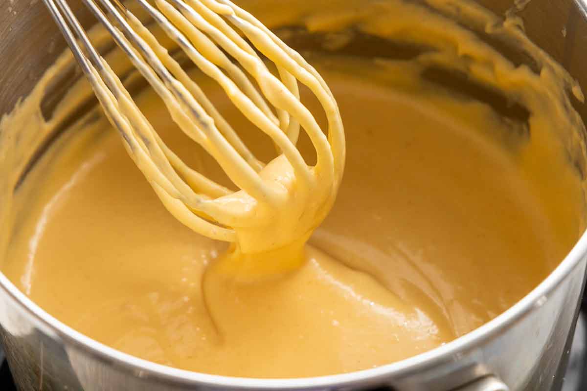 Whisk lifted from a pot showing melted cheddar fully incorporated into a smooth, creamy beer cheese sauce.