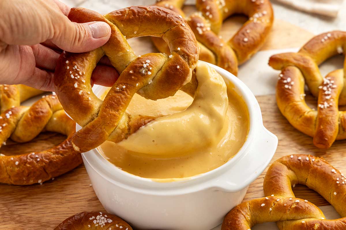 A soft pretzel being dipped into a bowl of smooth, creamy beer cheese dip, surrounded by more pretzels.