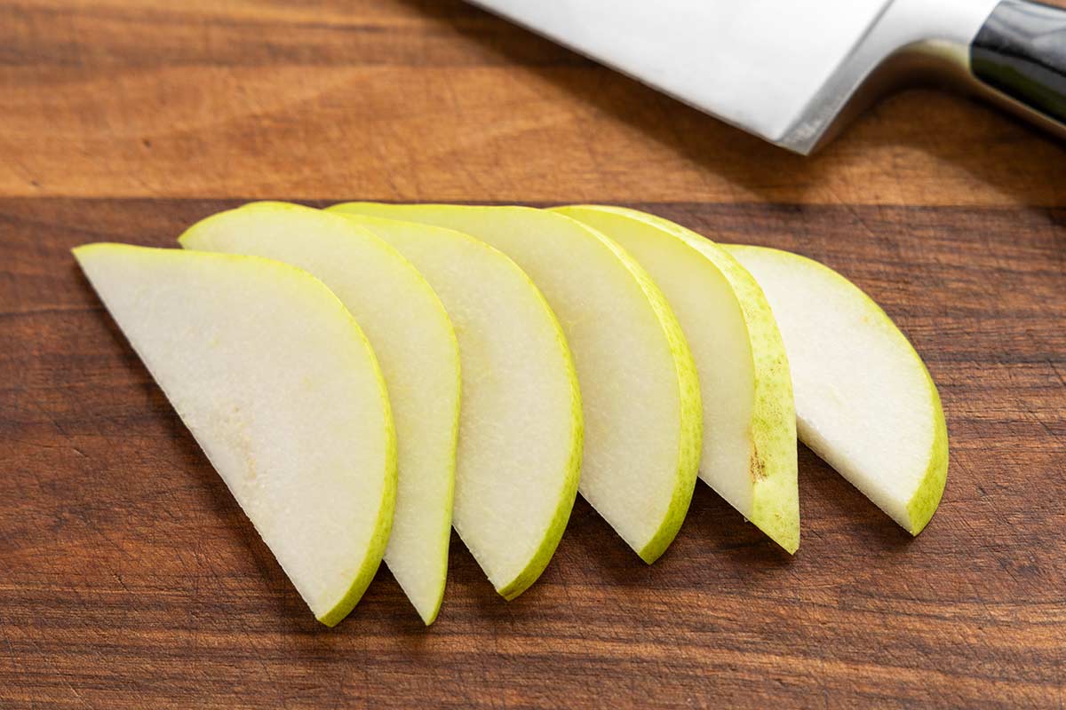 Thinly sliced green pear arranged on a wooden cutting board next to a chef's knife.