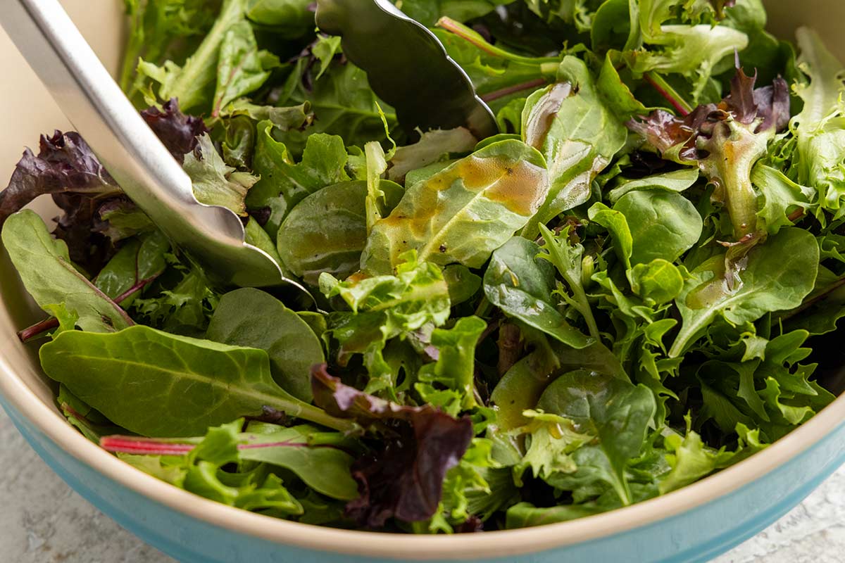 Mixed greens tossed with balsamic dressing in a bowl using metal tongs.